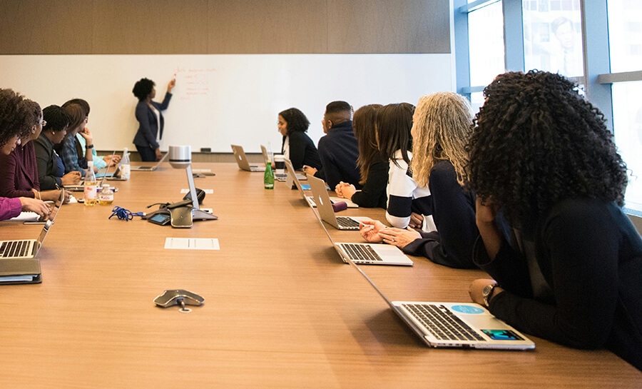 People in a meeting room with a woman leading a discussion at the whiteboard